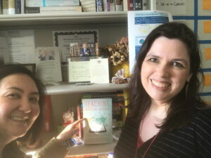 Two smiling women take a selfie in an office beside a bookshelf. The woman on the left points toward a displayed copy of Teaching Social Work with Digital Technology, while books, framed awards, papers, and office materials appear in the background.